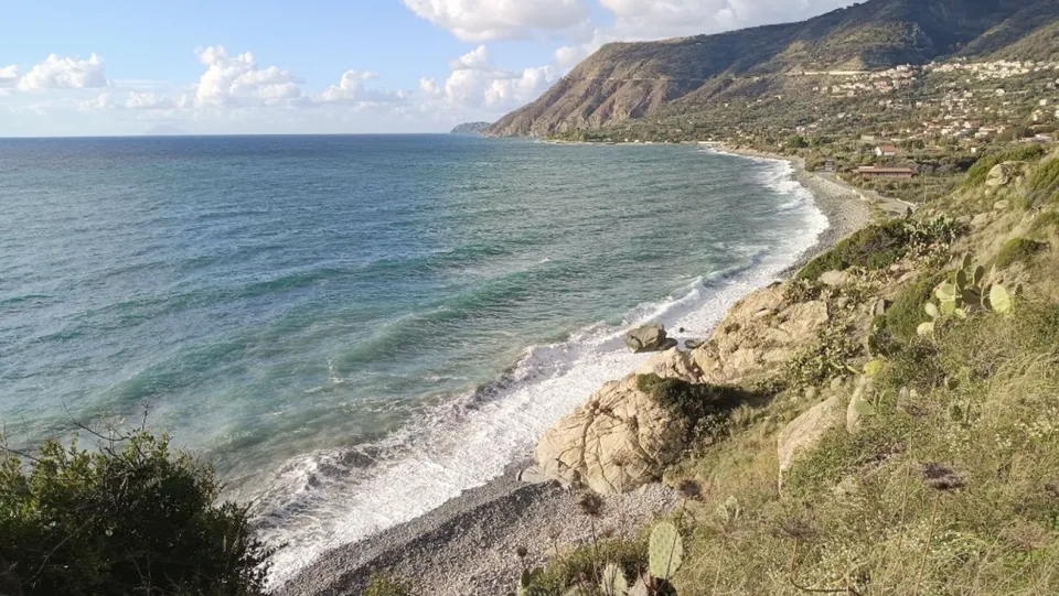 Kiesstrand mit Brandung, dahinter grüne Hänge und ein Ort an der Küste.