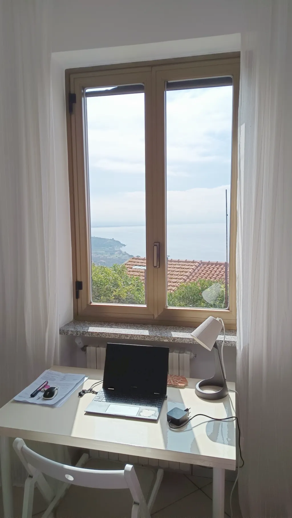 Ein weißer Schreibtisch mit Laptop vor dem geöffneten Fenster, draußen Dächer, die Küste und das Meer.